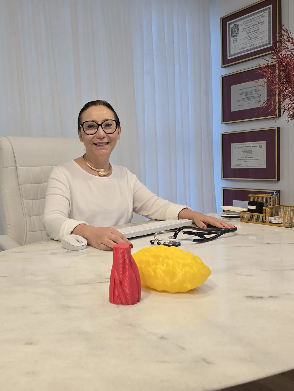 Dra. Divina Oliveira sorrindo na sala de consulta, sentada à mesa de mármore, com os diplomas emoldurados na parede ao fundo