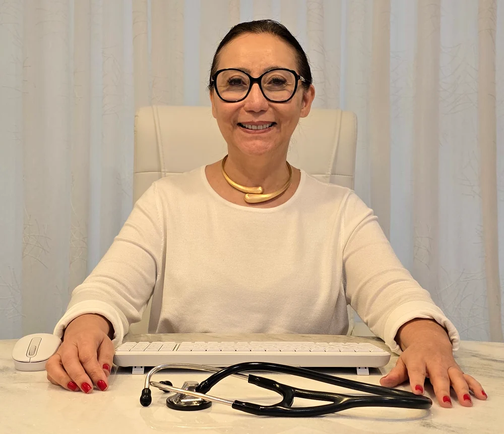 Dra. Divina Oliveira sorrindo em seu consultório, sentada à mesa com o estetoscópio